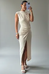 Vestido largo sin mangas de color beige, con cuello redondo y diseño drapeado en la cintura que crea un efecto de frunce. Presenta una abertura lateral pronunciada en la falda y un corte ajustado que estiliza la figura.