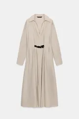 Vestido midi color beige claro, de manga larga y cuello solapa con escote pico. Presenta pliegues en la cintura y un cinturón negro contrastante con hebillas metálicas.