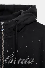 Sudadera negra con capucha, cierre frontal de cremallera metálica y mangas largas con puños. Presenta un diseño corto con bolsillo tipo canguro y detalle de texto 'California' aplicado con brillos en el frente.