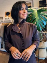 Camisa de color marrón oscuro con cuello mao y detalle de volados a lo largo de la abertura frontal. Presenta mangas largas con puños rectos y cierre frontal mediante botones ocultos.