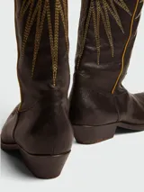 Botas de estilo cowboy de piel color marrón oscuro, con caña alta y punta afilada. Presentan bordados dorados en forma de estrella o rayos solares en la caña y un ribete lateral en hilo amarillo/dorado.