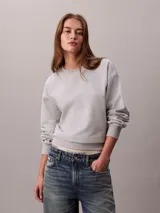 Buzo de felpa francesa gris claro, de cuello redondo y manga larga, con puños y dobladillo acanalados. Presenta un diseño holgado y lleva el logo retro de Calvin Klein bordado discretamente en el pecho.