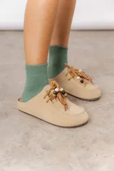Zueco de cuero gamuzado color tostado, con cordones decorativos adornados con cuentas de madera y piezas envueltas en cuero. Diseño tipo mocasín con costuras visibles en la parte superior y abierto en la parte trasera.
