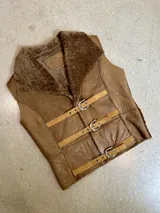 Chaleco de cuero genuino color marrón con interior de piel sintética. Presenta cuello solapa amplio y cierre frontal mediante tres correas ajustables con hebillas metálicas.