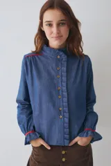 Camisa de denim azul con cuello alto tipo gorguera, cierre frontal con botones de madera y detalle de volados laterales. Presenta mangas largas con puños elásticos terminados en volados y detalles de costuras en color rojo en hombros y puños.