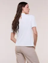 Blusa blanca de corte recto con cuello alto torzado, abertura frontal en gota y mangas cortas con aberturas sobre los hombros.