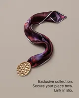 Pañuelo de seda con estampado multicolor en tonos violeta y burdeos, acompañado por un gran dije metálico dorado de textura martillada.