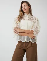 Blusa de tul bordado color beige, marca Liv Los Angeles, con cuello alto, cierre frontal de botones y mangas ¾ amplias con volados.