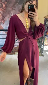 Vestido largo color bordó con escote en V profundo, detalle de nudo central y aberturas laterales en la cintura. Presenta mangas largas abullonadas con puños elásticos y falda con tajo frontal pronunciado.