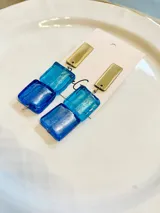 Par de caravanas colgantes con piezas rectangulares de vidrio azul y celeste, y piezas rectangulares doradas en la parte superior.