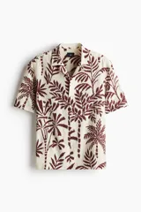 Camisa de manga corta con cuello abierto, cierre frontal de botones y bajo recto. Presenta un estampado de palmeras en color marrón sobre fondo beige.