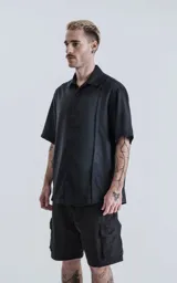 Camisa de manga corta en color negro con proceso de lavado, cuello clásico y cierre frontal con botones. Presenta un diseño minimalista con una costura vertical decorativa en el frente.