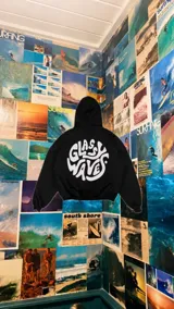 Hoodie oversize negro con capucha y bolsillo tipo canguro, confeccionado con un gran estampado circular blanco en la espalda que presenta la inscripción "Glassy Waves" en tipografía fluida y psicodélica.