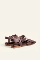 Sandalia de cuero negro estilo huarache, con puntera cerrada mediante tiras finas entretejidas a mano, tira ajustable al tobillo con hebilla metálica y suela plana con taco de 1 cm.