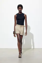 Bermuda color crema de tiro medio con pinzas delanteras y cinturón fino color beige. Tiene bolsillos frontales y falsos bolsillos de vivo en la espalda. Cierre frontal con cremallera, botón interior y gancho.