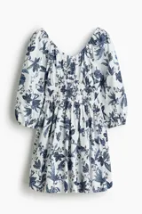 Vestido corto de algodón con estampado floral en tonos azules sobre base blanca, escote en V y mangas globo de longitud al codo. Presenta elástico fruncido en el pecho y la cintura, con falda evasé de vuelo.