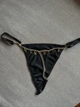 Tanga negra con tiras regulables y detalles de cadena dorada con dijes colgantes que forman la palabra 'BANDIDA'.