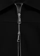 Chaqueta negra de corte recto con cuello camisero y cierre frontal de cremallera doble metálica. Presenta diseño de manga larga y estructura entallada.