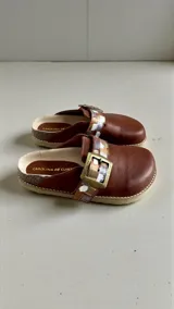 Sandalias de cuero color marrón de la marca Carolina de Cunto, modelo SAKI. Presentan una tira con hebilla metálica grande y detalles pintados a mano en tonos dorados, plateados y blancos. Cuentan con diseño destalonado y suela plana.