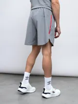 Short de entrenamiento Umbro modelo Grit, color gris, confeccionado en tejido fourway stretch. Presenta detalles en contraste rojo, incluyendo una franja lateral vertical y el logo Umbro bordado en el muslo izquierdo. Posee cordón de ajuste en la cintura y un bolsillo lateral con cierre.