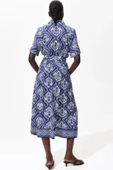 Vestido midi camisero de color azul con estampado ornamental en tonos blancos y azules. Cuenta con mangas cortas raglán, botonadura frontal completa y cinturón de la misma tela para atar en la cintura.