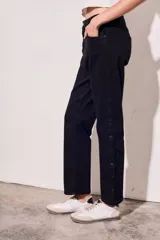 Pantalón de jean negro, de corte recto y tiro medio. Presenta bolsillos delanteros y traseros, y una hilera de botones metálicos a lo largo de la costura lateral.