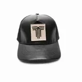 Gorra tipo trucker de color negro, con frente de cuero sintético y parte trasera de red. Presenta un parche rectangular en el centro con el logo de la marca bordado en color negro sobre fondo beige.