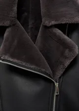 Chaqueta estilo biker de efecto piel en color negro, con cuello y forro interior de pelo sintético en tono gris oscuro. Presenta cierre frontal de cremallera, manga larga y diseño estructurado.