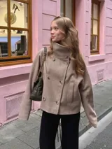 Chaqueta corta de paño en color beige, con cuello alto, cierre frontal cruzado de doble botonadura y mangas largas holgadas.