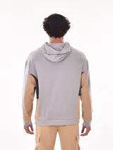 Buzo canguro negro con capucha, diseño de bloques de color en negro, beige y gris claro en el torso y mangas. Presenta un pequeño detalle de texto blanco en el pecho.