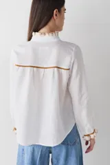 Camisa de lino blanca con detalles de volados tejidos a mano en crochet en cuello, cartera y puños. Presenta vivos a contratono en color marrón y cierre frontal con botones.
