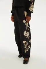 Pantalón de jogging negro de tiro medio, con cintura elastizada y cordón ajustable. Presenta un diseño holgado y estampado botánico localizado en tono claro sobre una de las piernas.