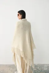Ruana de punto color crudo, confeccionada en lana merino sin teñir con mezcla de lino y viscosa, con diseño triangular y terminación de flecos en los bordes.