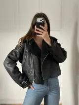Campera tipo gamulán de cuero sintético negro con interior y cuello forrados en corderito sintético. Presenta cierre frontal cruzado, bolsillos laterales con cierre y correas ajustables con hebilla en los puños y en el ruedo.