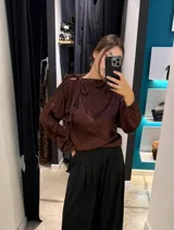 Blusa de satén color negro con cuello alto, mangas largas con detalle de botones laterales y lazo decorativo en el hombro.