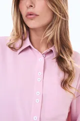 Camisa blanca de manga larga con cuello clásico y cierre frontal de botones negros en contraste. Presenta un bolsillo frontal en el pecho y un diseño de líneas simples y corte recto.