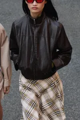 Chaqueta estilo bomber corta de efecto piel en color marrón oscuro, con cuello subido, cierre frontal con cremallera y cintura elástica fruncida.