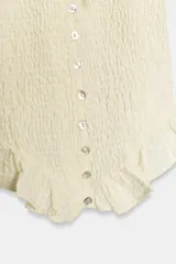 Top color crema con cuello redondo y lazada, escote en pico y mangas cortas abullonadas con elástico. Presenta un tejido elástico tipo nido de abeja y cierre frontal con botones.