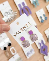 Aros colgantes de diseño geométrico, compuestos por una pieza cuadrada superior esmaltada en color lila y una pieza cuadrada inferior decorada con cristales brillantes en tonos violeta.