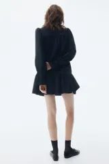 Vestido corto de corte holgado con cuello alto de volados y cierre frontal de botón. Presenta canesú fruncido, mangas largas abullonadas con puños acampanados y falda con costura fruncida en el bajo.