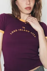 Camiseta de cuello redondo y manga corta, color azul marino con ribetes en color rojo en cuello y mangas. Presenta un estampado frontal ovalado con el texto New York en color naranja.