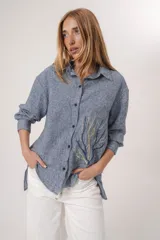 Camisa de lino azul con textura jaspeada, corte holgado y manga larga. Presenta un bordado decorativo en el frente que simula una rama o coral en tonos amarillos y azules, con cierre frontal de botones.