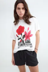 Remera de corte oversize y silueta boxy, color blanco, con cuello redondo y mangas cortas. Presenta una estampa frontal con diseño de flores en tonos rojos y rosados con efecto spray y tipografía negra.