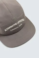 Gorra de seis paneles color marrón topo con visera plana y ojales bordados. Presenta bordado frontal en blanco con el texto "Alternative Lifestyle" y "Trastevere, Rome.".