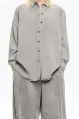 Camisa de manga larga color gris claro con efecto lavado, corte recto y botones frontales marrones. Presenta cuello camisero y pespuntes a contraste.