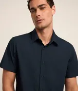 Camisa de manga corta con cuello inglés y cierre frontal abotonado, confeccionada en una mezcla de fibras de bambú, viscosa, poliéster y elastano. Presenta un diseño liso y entallado.