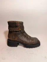 Bota corta de cuero vacuno con textura desgastada en tono marrón oscuro. Presenta una suela de goma tipo track, cierre lateral interno y una tira decorativa con hebilla metálica en el tobillo. Diseño de punta cuadrada.