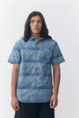 Camisa de manga corta Quiksilver modelo Alan, con estampado geométrico en tonos azules, lavanda y beige. Presenta cuello camisero, cierre frontal de botones y un bolsillo aplicado en el pecho con detalle de logo.