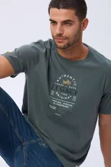 Remera T-shirt de algodón color verde oscuro, con estampado frontal en blanco y amarillo que incluye texto "Authentic Legacy The Original 1972" y el dibujo de una motocicleta. Tiene un pequeño detalle de etiqueta marrón en la manga.