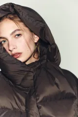 Campera puffer corta de color marrón oscuro, con diseño acolchado, cuello alto y capucha integrada. Presenta cierre frontal con botones magnéticos y mangas largas con puños elásticos.